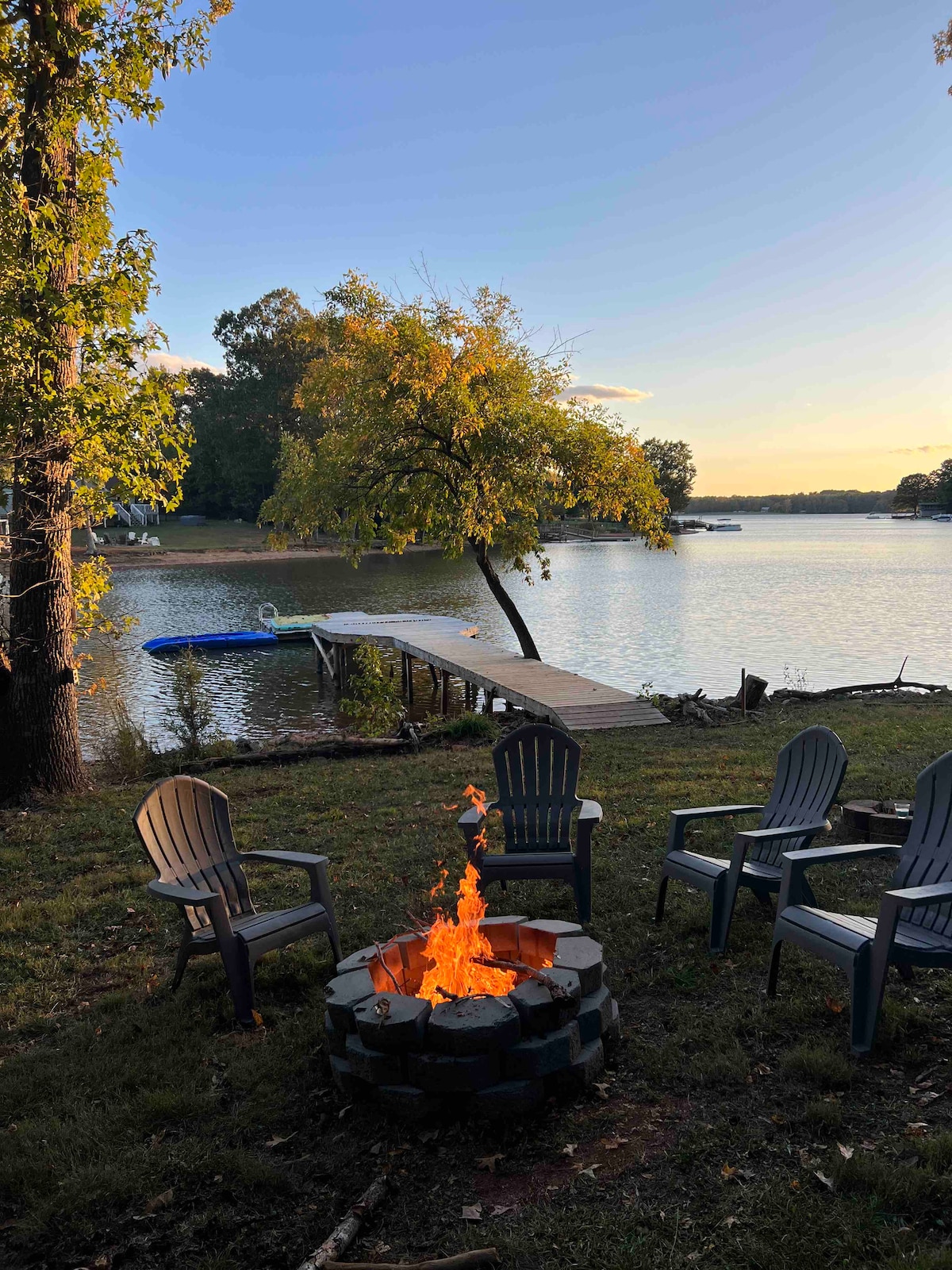 A circular stone fire pit surrounded by several Adirondack chairs is shown in a serene outdoor setting. A private dock extends into the calm water, framed by trees and the soft glow of sunset illuminating the scene.