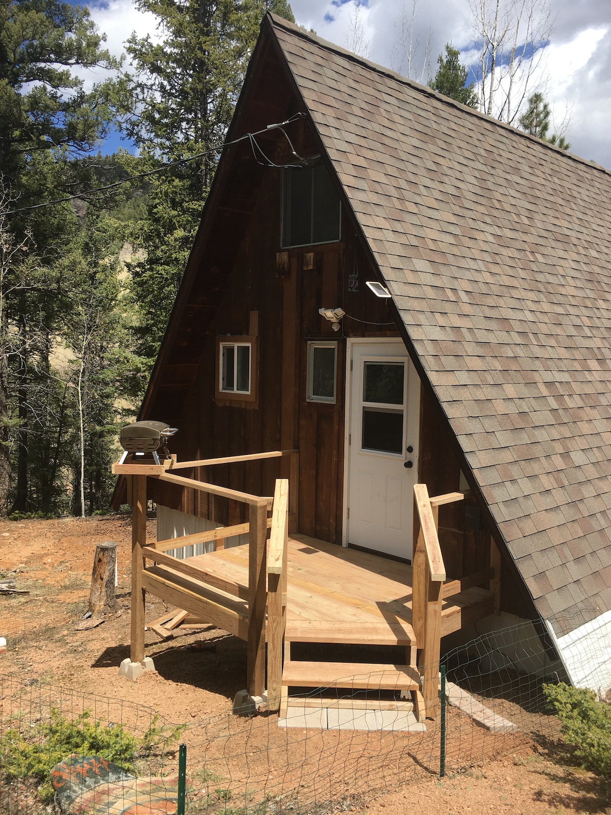 An A-frame cabin is set amidst tall pine trees, featuring a wooden front deck with steps leading to the entrance. The natural surroundings and rustic wood exterior reflect a cozy mountain retreat.