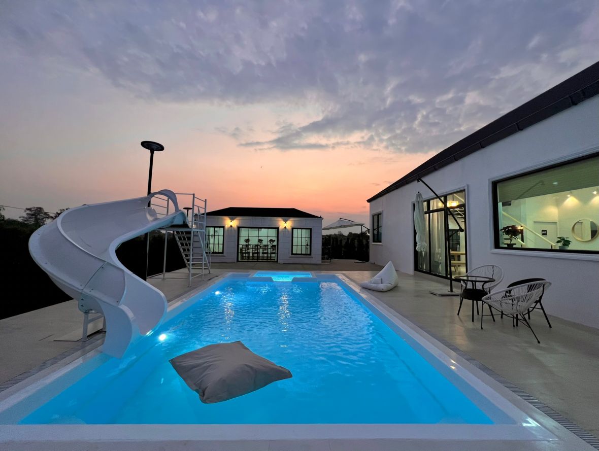 A private swimming pool designed with a water slide is visible, surrounded by a spacious patio. Soft seating areas are provided, with a large sky illuminated by sunset hues. The villa's large windows reflect the warm light, creating a welcoming outdoor space.