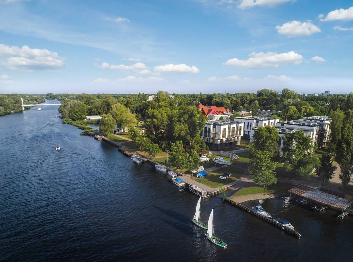 Aerial view of a serene river bordered by lush greenery and a variety of boats. Two sailboats glide across the water, while several moored vessels line the shore. The apartment complex is situated among trees, offering a natural backdrop to the waterside setting.