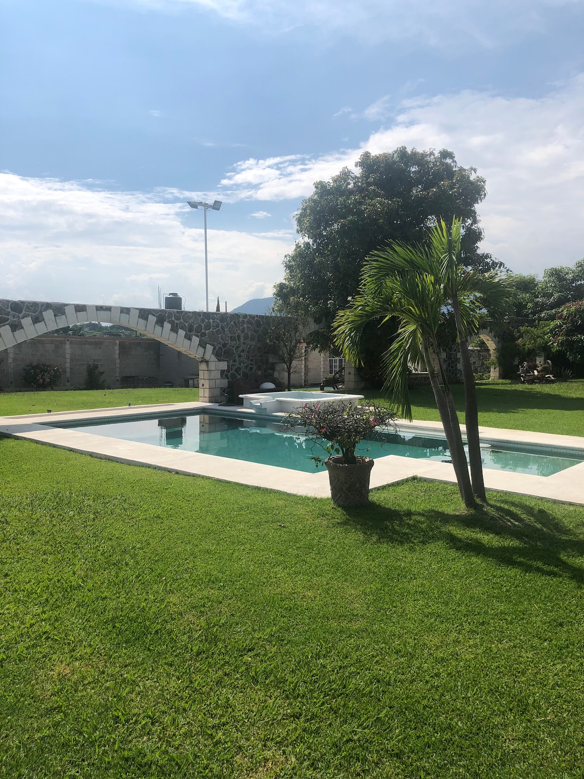 A serene outdoor space features a clear swimming pool surrounded by a well-maintained lawn. Lush greenery, including tall palm trees, provides shade. A stone archway adds architectural interest in the background, complementing the tranquil atmosphere of the area.