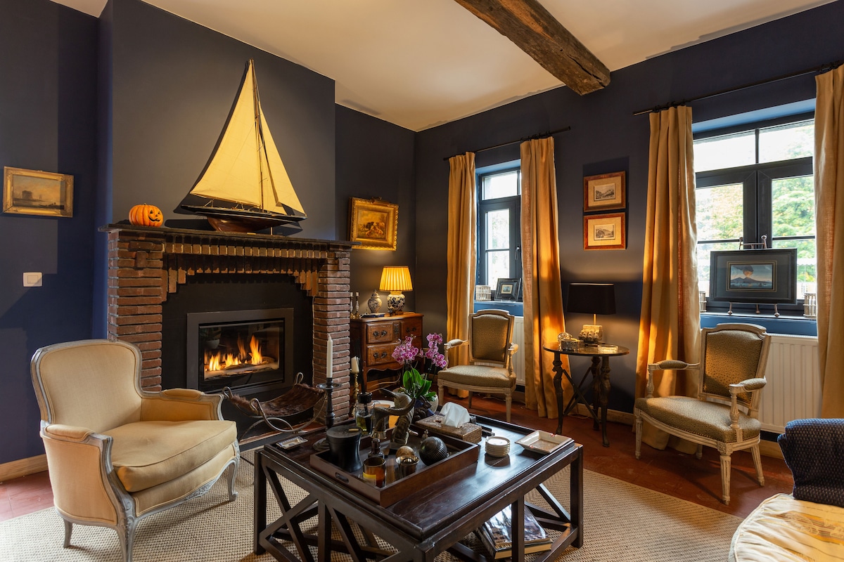 A comfortable living area is presented, featuring a brick fireplace with a decorative sailboat above it. Two upholstered chairs are positioned beside a dark wood coffee table, which holds various decorative items. Windows with dark frames allow natural light to fill the space.