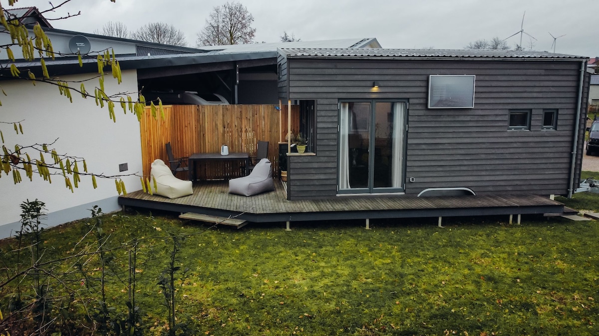An exterior view of a modern tiny house is presented, showcasing a wooden deck with seating options and a small table. The structure features large glass doors and large windows, while a surrounding grassy area adds to the natural setting. A wooden fence provides privacy.