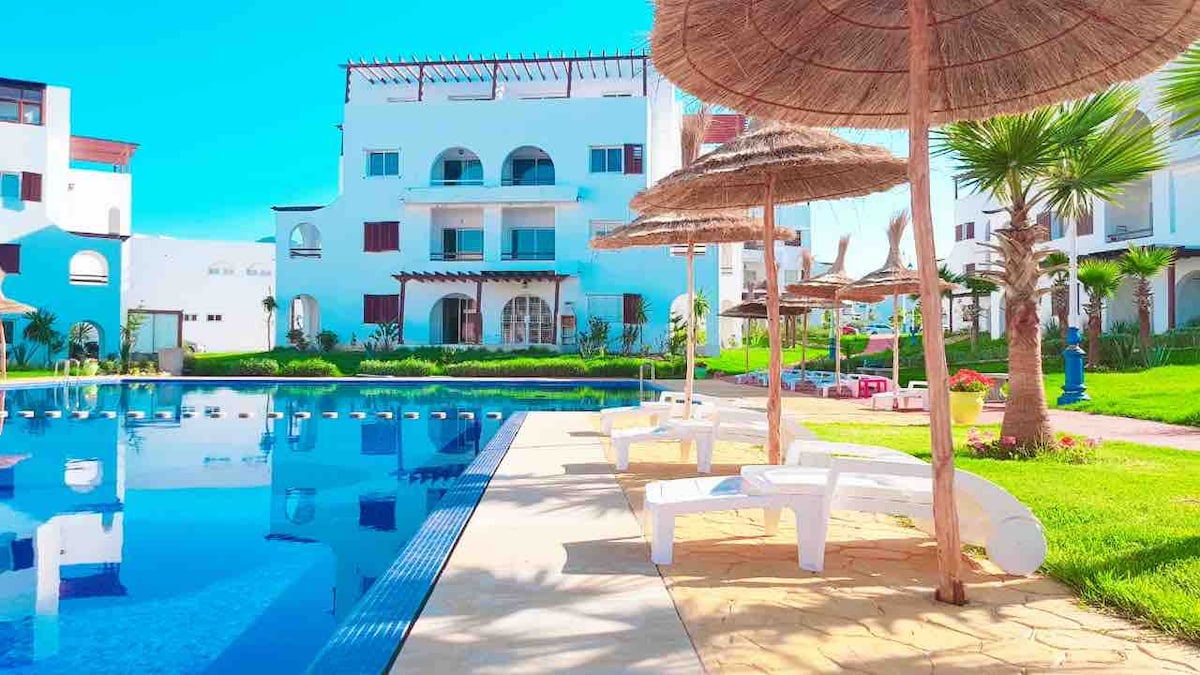 A swimming pool reflects the clear blue sky, framed by lounge chairs and straw umbrellas. Lush greenery surrounds the area, complementing the modern buildings in the background, which feature arched windows and balconies, offering a serene atmosphere for relaxation.