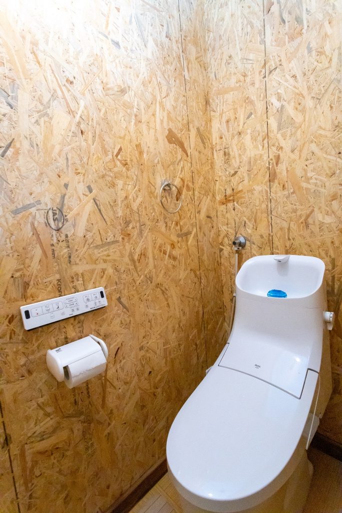 A modern toilet with an integrated washlet feature is positioned against a wall made of oriented strand board. A toilet paper holder is affixed to the wall nearby. The interior reflects a clean and functional design.