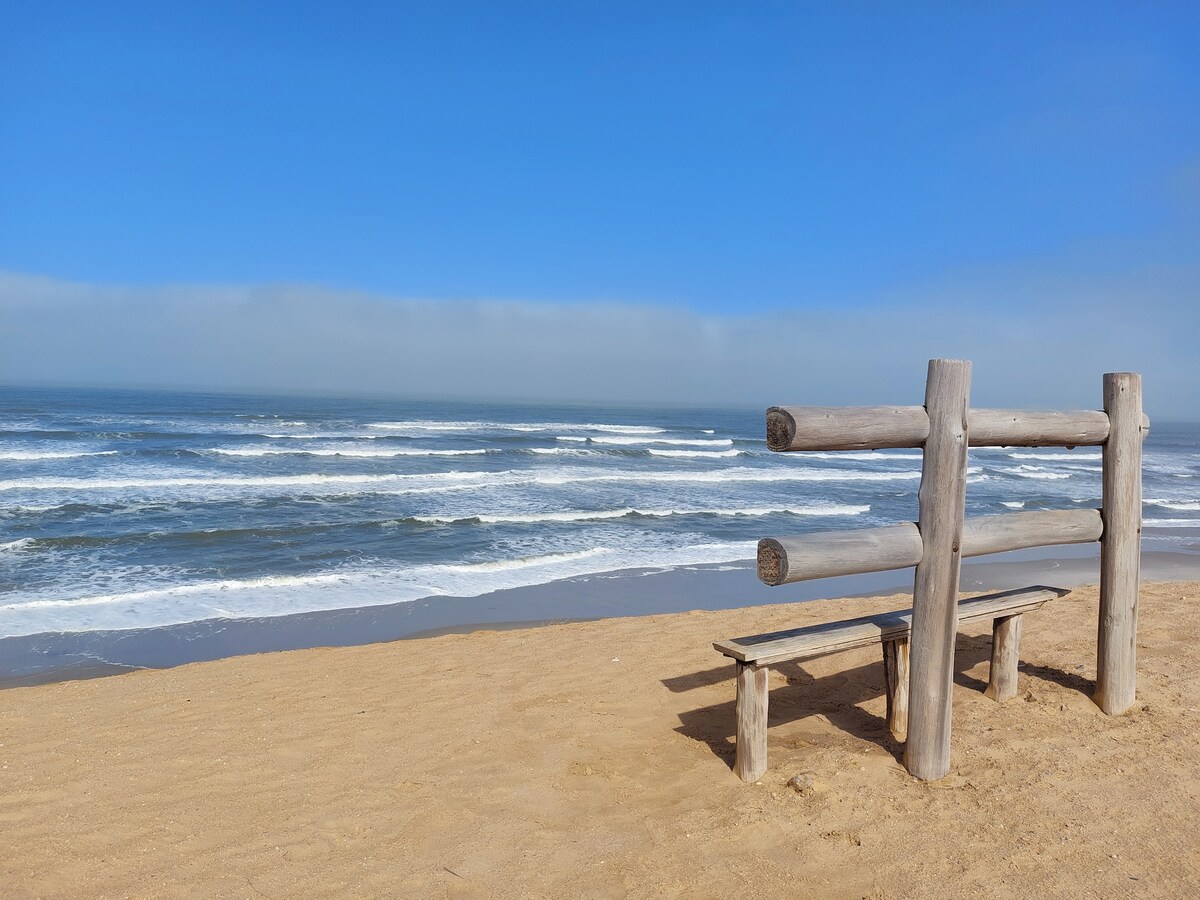 A wooden bench and railing are positioned on a sandy beach, overlooking rolling waves gently lapping the shoreline. The clear blue sky contrasts with a soft haze on the horizon, creating a serene coastal scene.