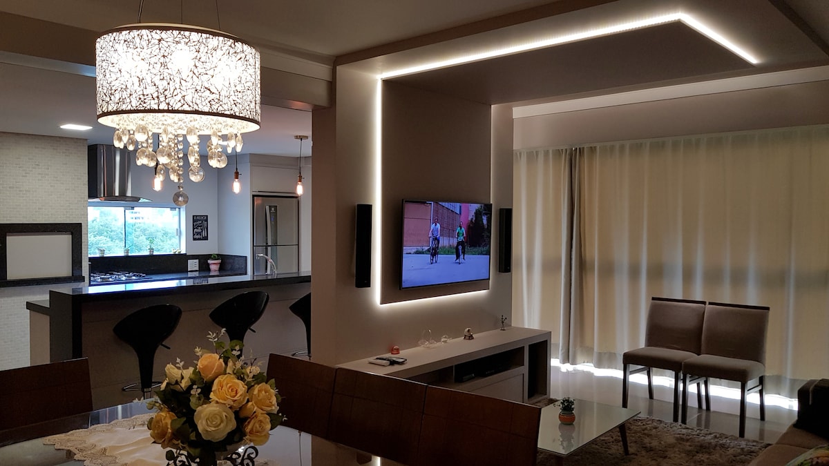 A contemporary living area features a stylish chandelier with crystals, illuminating the space. A large television is mounted on the wall opposite two elegant chairs and a dining table adorned with a bouquet of yellow roses. Soft curtains diffuse natural light throughout the room.
