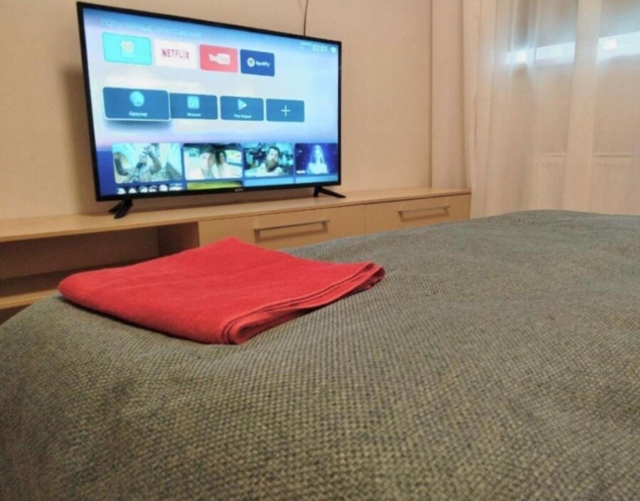 A neatly made bed is presented with a textured blue blanket, complemented by a folded red towel. A large flat-screen television is mounted on the wall, displaying a user-friendly interface with icons for entertainment options below a window covered by sheer curtains.