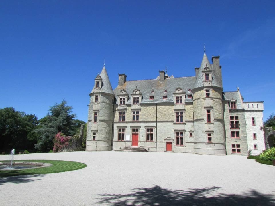 A grand stone castle is depicted, featuring elegant turrets and large windows with red accents. The well-maintained grounds include a circular flowerbed and are bordered by lush greenery, creating a serene backdrop against the clear blue sky.