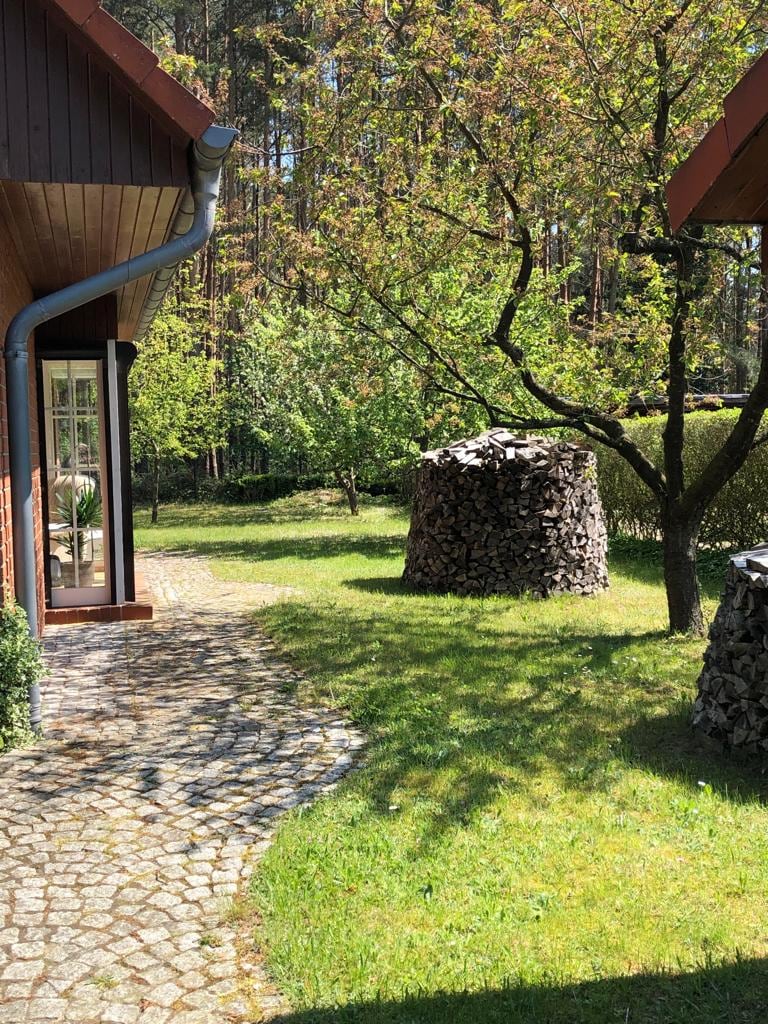 A stone pathway winds through a lush green garden, bordered by trees and shrubs. A round stack of firewood is neatly arranged nearby, and another tree adds to the serene atmosphere, offering a glimpse of nature's tranquility.