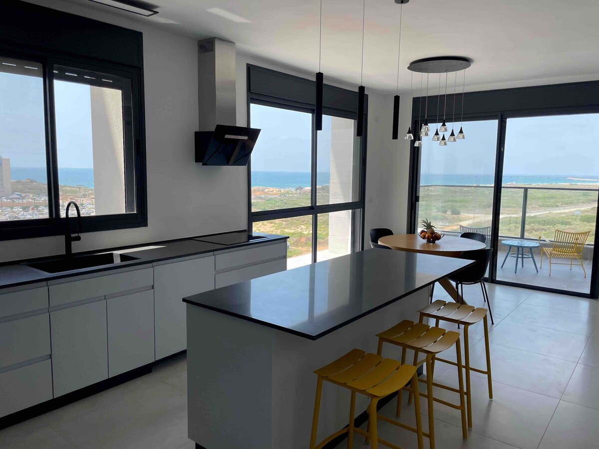 A modern kitchen is showcased with sleek cabinetry and a large island featuring bar seating. Floor-to-ceiling windows provide abundant natural light and an expansive view of the sea and surrounding landscape. A dining area with stylish chairs is visible in the background.