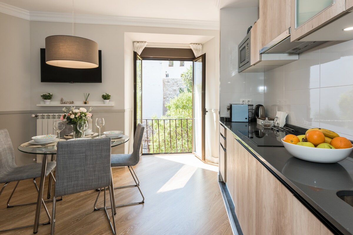 A modern kitchen area is visible with sleek cabinetry and a black countertop. A round dining table is set for four, featuring elegant glassware and a vase of flowers. Large windows offer natural light and a view of greenery outside, enhancing the inviting atmosphere.