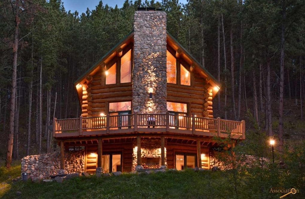 The custom log home features a striking stone chimney and large windows that reflect the surrounding forest. A spacious deck wraps around the home, providing a connection to nature. Subtle outdoor lighting enhances the inviting appearance during dusk.