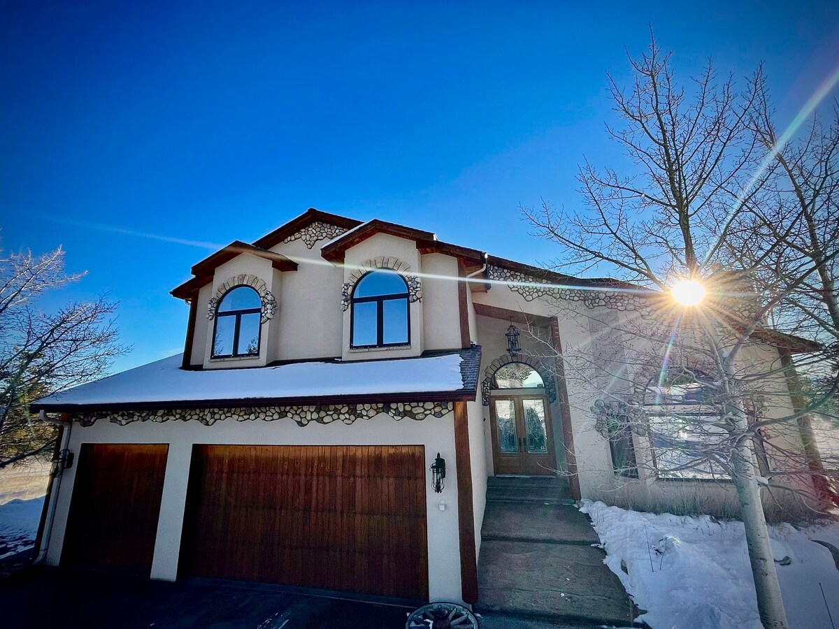 A large, two-story house features a symmetrical facade with arching windows and a welcoming entry. Snow covers the roof and driveway, while trees frame the property. A sunburst highlights the scene, creating a serene winter atmosphere.