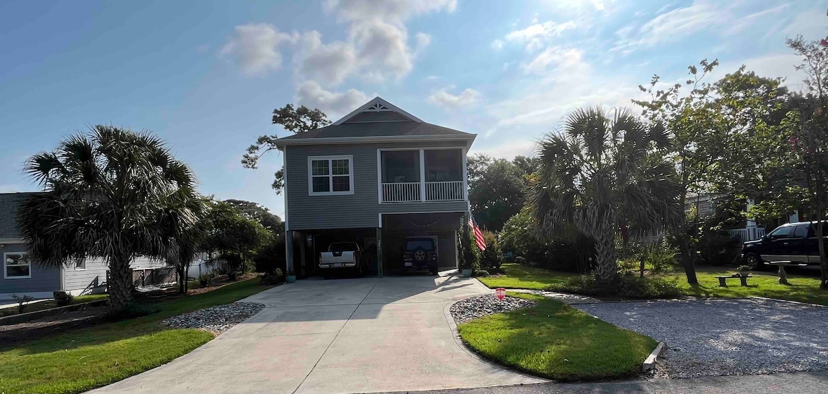 A two-story house is presented with a wide concrete driveway and landscaped lawn. Palm trees cluster around the property, complementing the inviting exterior. A covered porch extends over the entrance, and a flag hangs cheerfully from the balcony. Clear skies enhance the home's natural lighting.