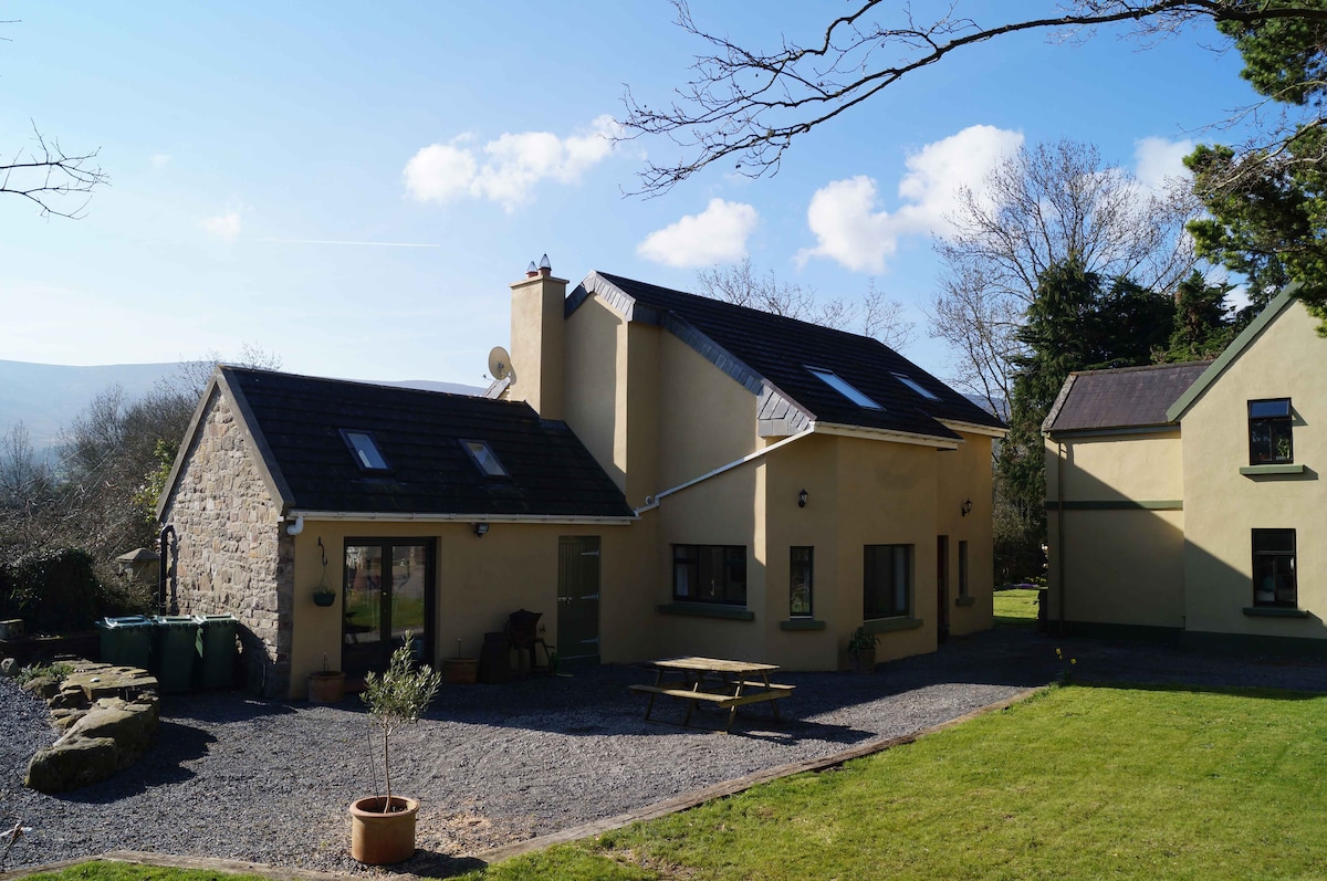 A cozy stone and stucco cottage is set against a clear blue sky, surrounded by green lawns and trees. A gravel patio area features a picnic table, inviting outdoor relaxation, while large windows allow natural light into the inviting living spaces.