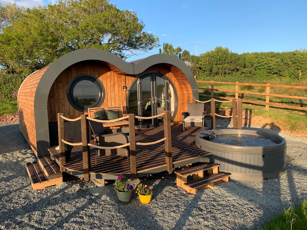 The exterior of The Willow features a unique curved design with large circular windows. A spacious deck is equipped with outdoor seating and a hot tub, surrounded by gravel and planters with flowers. Lush greenery and a wooden fence create a serene backdrop.