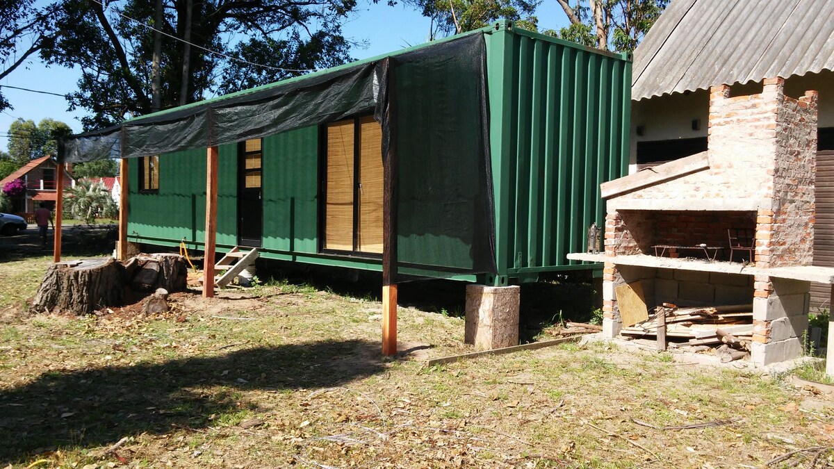 A green shipping container is positioned in the landscape, featuring large windows with wooden frames. Adjacent to the container, a brick barbecue area is visible with a stone base, and a cut tree stump lies nearby, set against a backdrop of grass and trees.