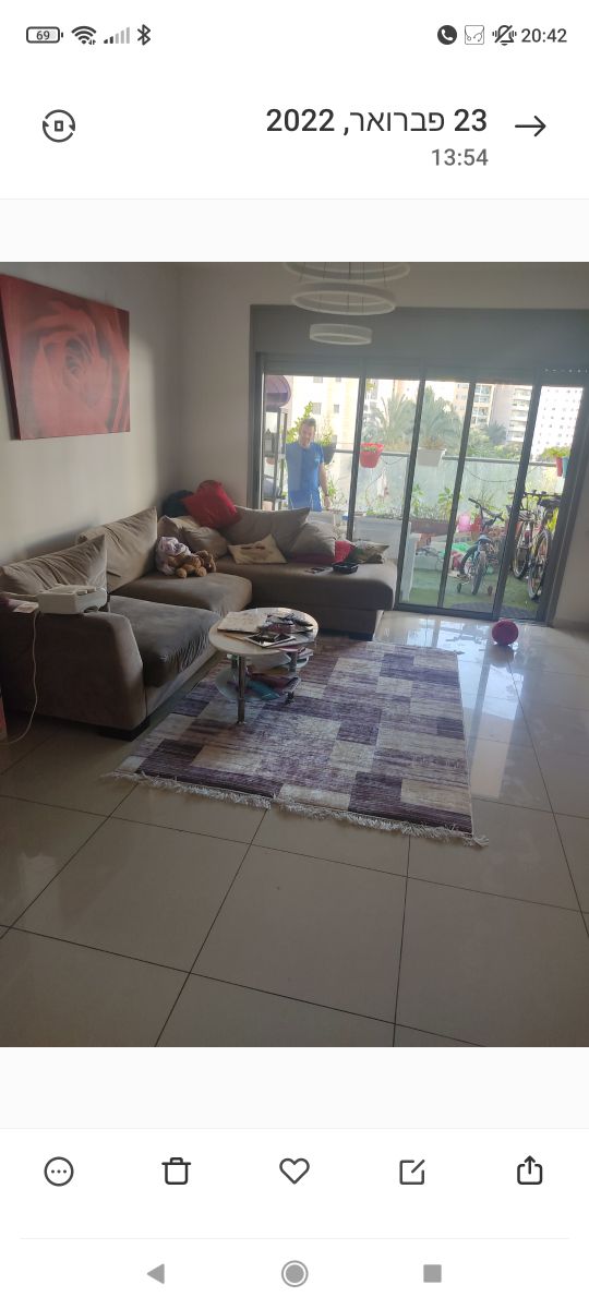 A living area features a large, comfortable sofa adorned with colorful cushions. A glass coffee table sits on a patterned rug, and a bright outdoor view can be seen through sliding glass doors. Natural light fills the space, enhancing its open feel.