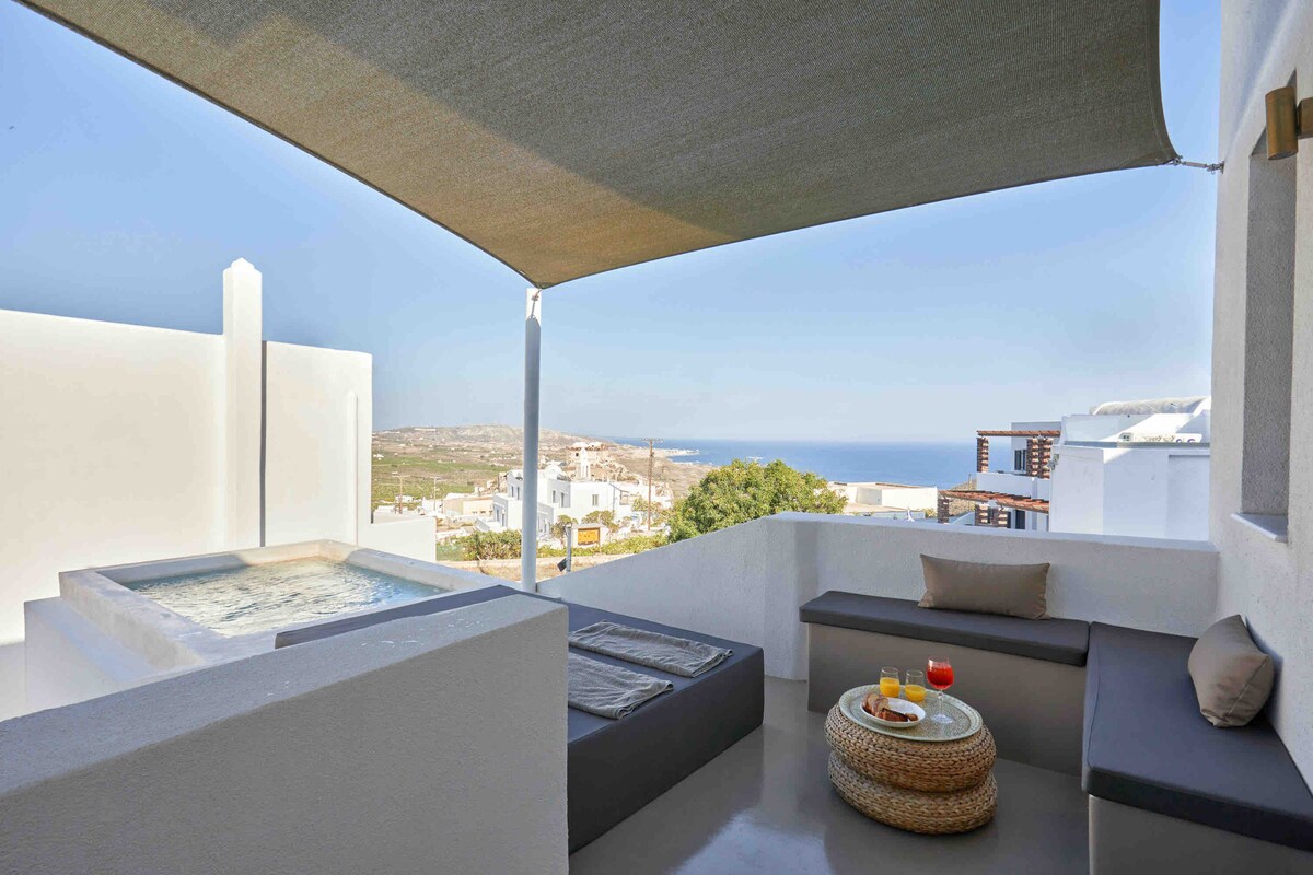 A shaded terrace features a private jacuzzi surrounded by sleek seating. A low table is adorned with fresh drinks, with expansive views of the sea and surrounding landscape visible in the background.