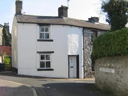 The exterior of Albion Cottage is presented, featuring a quaint two-story structure with a whitewashed facade and stone accents. A black door is visible at the front, framed by a neatly maintained hedge. A street sign indicates the location on Back York Street.