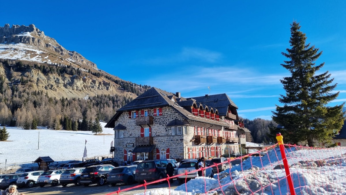 A rustic building with a stone facade and red accents is framed by snow-covered slopes and mountains. The structure features multiple balconies and large windows. A parking area is visible in front, surrounded by a red safety barrier. Clear blue skies enhance the picturesque setting.
