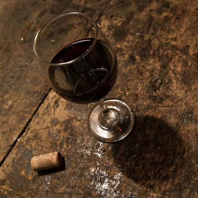 A clear wine glass filled with red wine is positioned on a rustic wooden surface. A cork rests alongside the glass, highlighting the natural textures of the table. Soft lighting casts a gentle shadow, creating a warm and inviting detail.