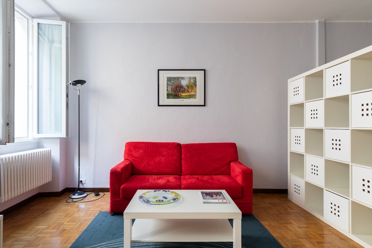 A living area features a vibrant red sofa paired with a white coffee table. A framed artwork hangs on the wall above the table, while natural light enters through a large window. A tall lamp stands next to the sofa, and a bookshelf is positioned against the opposite wall.
