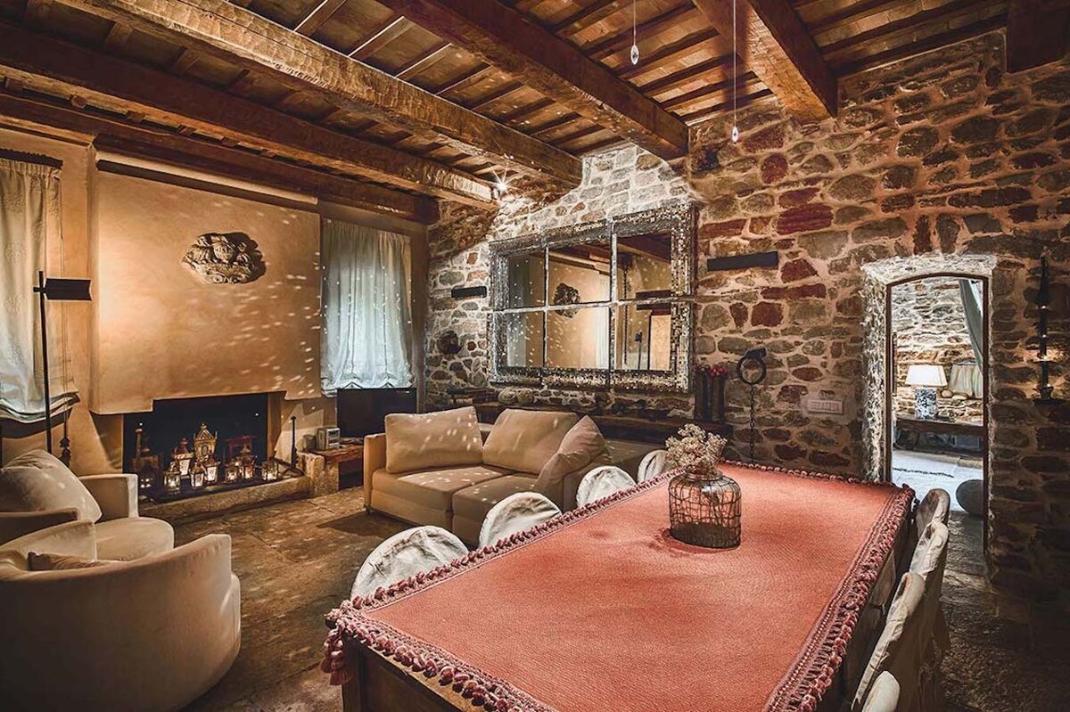 A spacious living area is presented, featuring a rustic stone wall and wooden beams overhead. Comfortable seating is arranged around a large table with a decorative centerpiece. Subtle lighting creates a warm ambiance, while a mirror reflects the inviting atmosphere of the space.