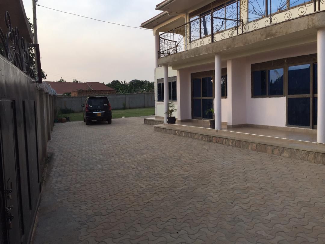 A spacious driveway is presented, paved with patterned stones, leading up to a two-story house. The exterior features large windows that allow for ample natural light. Surrounding greenery adds a sense of openness, while a parked vehicle is visible along the left side.