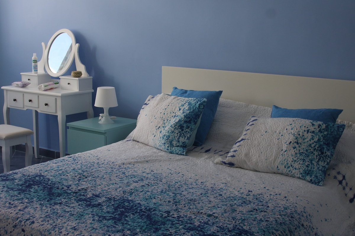 A serene bedroom is presented, featuring a spacious bed adorned with textured blue and white linens. A wooden bedside table with a lamp stands next to the bed, while a vanity with a round mirror is visible along the wall, complemented by a coordinating chair.