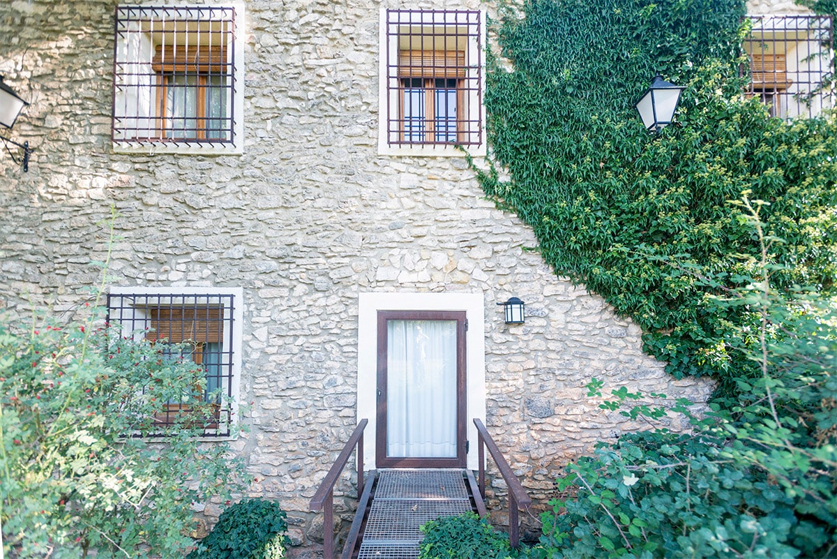 The stone facade of the building displays an earthy texture, with greenery climbing the walls. A wooden door is framed by a small wooden ramp leading to it, while four windows, protected by iron bars, add character to the exterior.