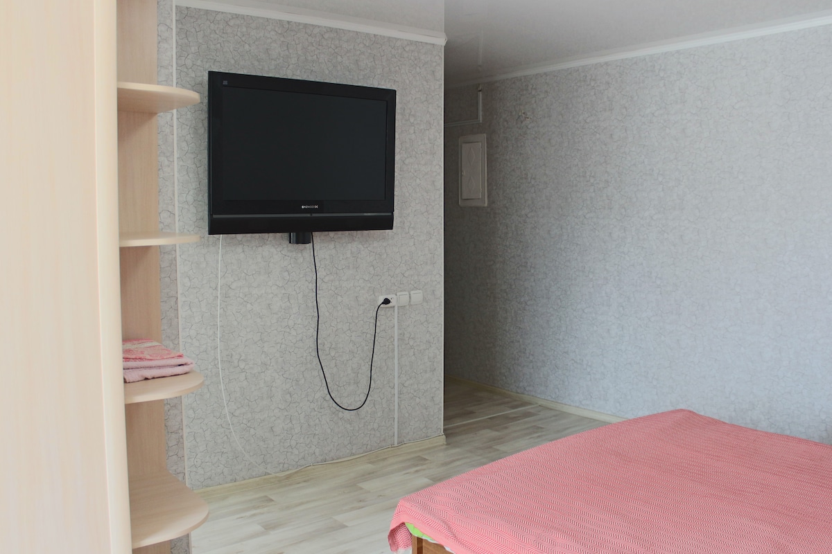 A bedroom is shown featuring a spacious bed covered with a soft pink blanket. A large television is mounted on the wall, with a cable running down. Light-colored flooring contrasts with light grey patterned walls, creating a subtle and relaxed atmosphere.