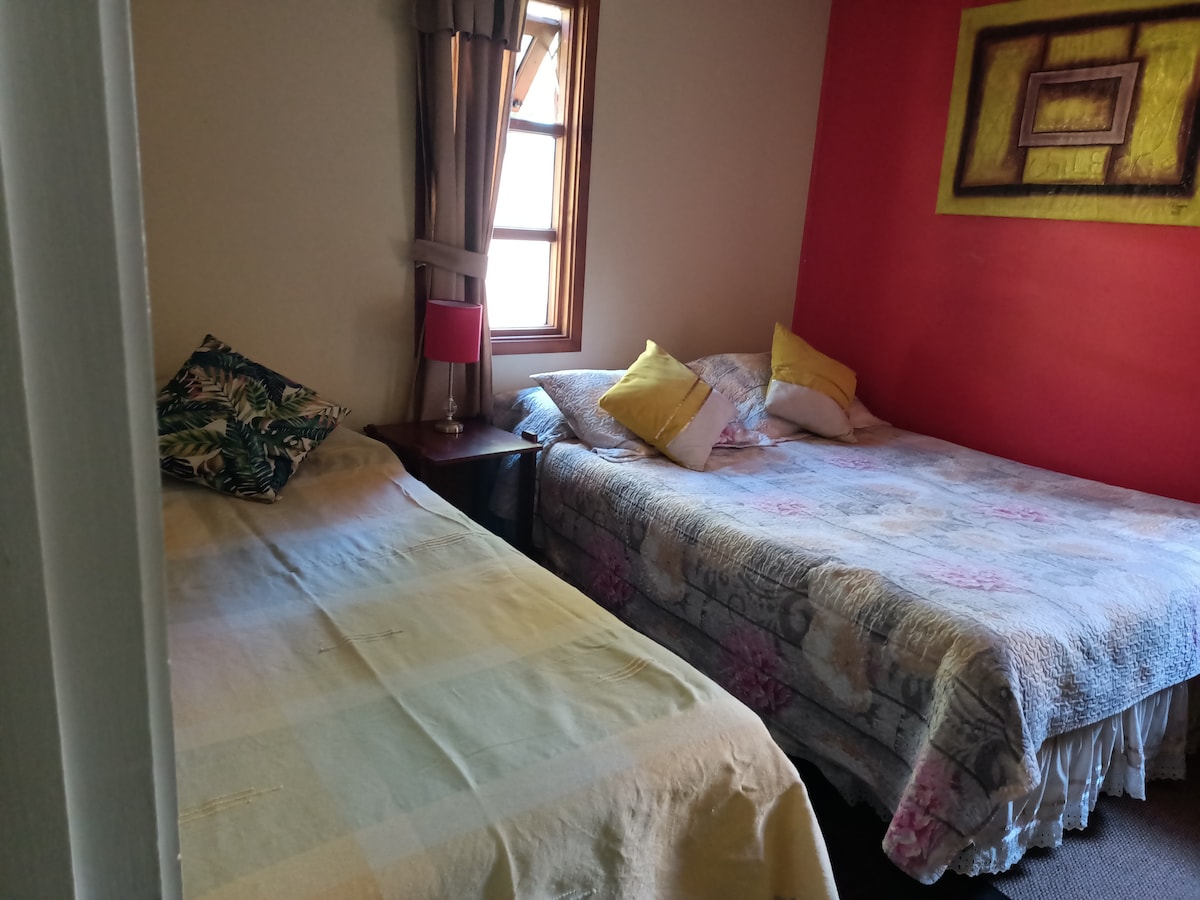 A sleeping area contains two spacious beds, each adorned with patterned bedding. A small bedside table with a lamp is positioned between the beds. Natural light enters through a window, illuminating the warmly colored walls.