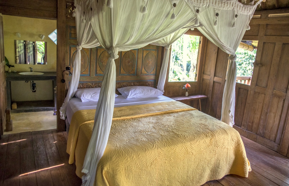 A cozy bedroom features a spacious bed dressed in a yellow coverlet, surrounded by natural wood paneling. Flowing white drapes add a soft touch. The adjoining bathroom is visible, showcasing modern fixtures. Natural light filters in, enhancing the serene setting amidst lush greenery.