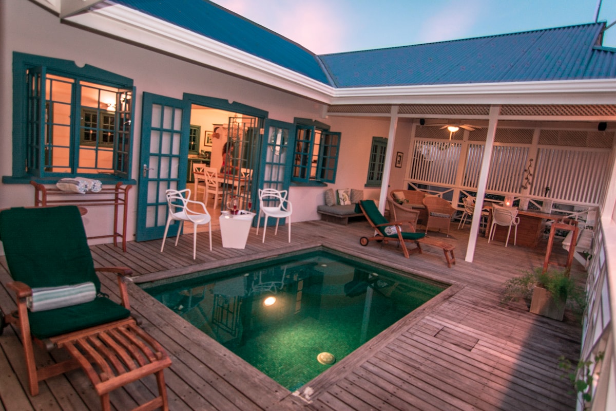 A serene outdoor space features a small, inviting pool surrounded by wooden decking. Comfortable lounge chairs are arranged for relaxation, while a dining area is visible in the background. Multiple sets of blue-framed French doors open to create a seamless connection between indoor and outdoor living.