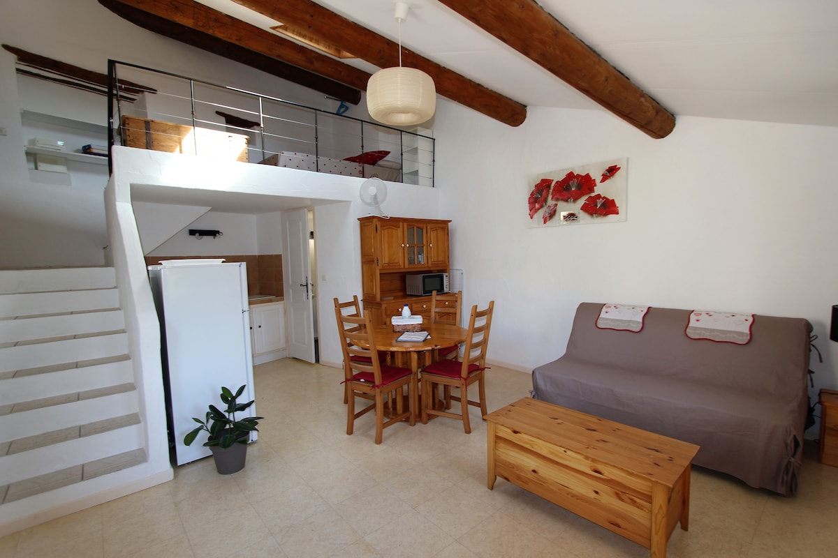 A spacious interior is displayed, featuring a cozy living area with a sofa and wooden coffee table. A dining table with chairs is positioned nearby. An open staircase leads to a mezzanine level above, while a kitchenette is visible in the background. Light enters through the windows.