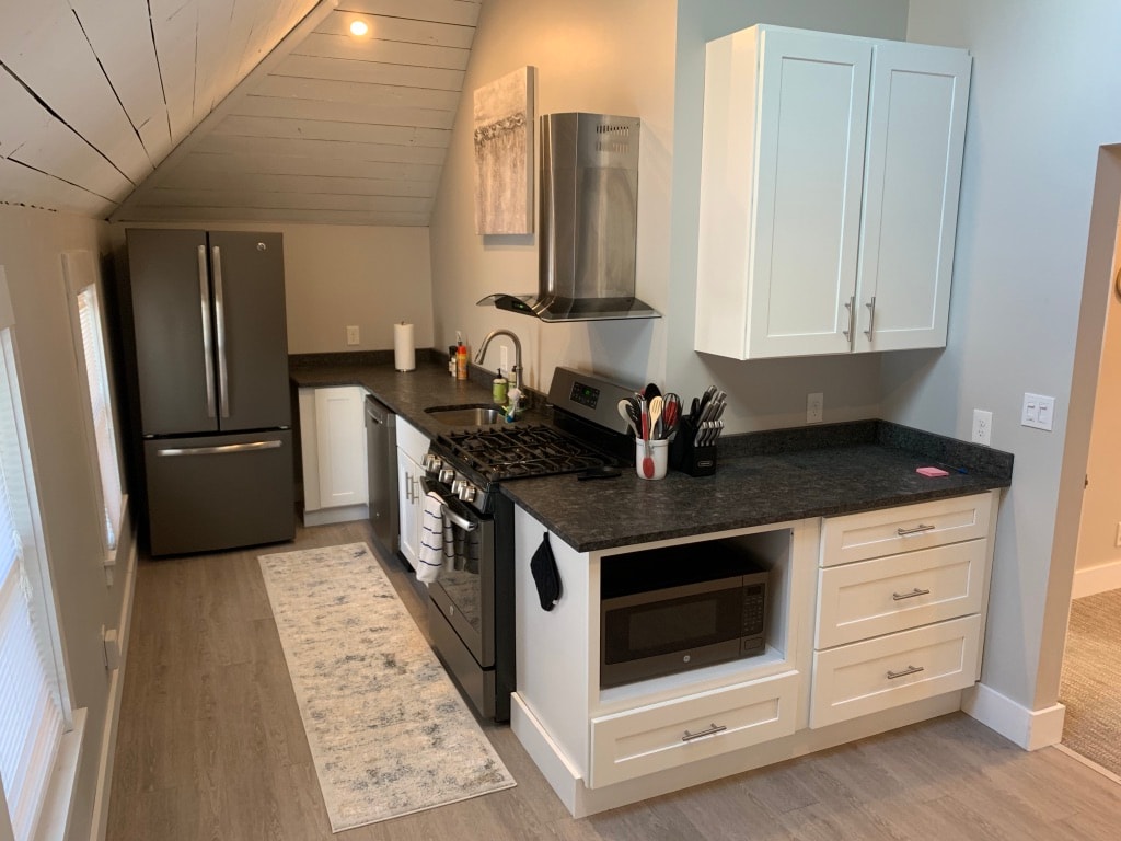 A modern kitchen features sleek white cabinetry and a dark countertop. A stainless steel refrigerator and gas stove are visible, alongside a microwave. Natural light enters through nearby windows, illuminating the space. A patterned rug lies on the floor, adding a touch of comfort.