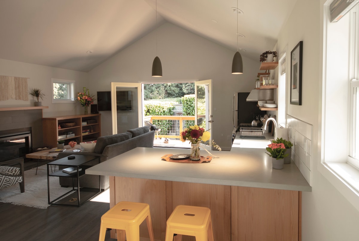 A bright, open kitchen and living area features a centrally located counter with two yellow stools. Large glass doors open to a private backyard, complemented by modern decor and natural light. A variety of plants and flowers are arranged on the counter, adding color to the space.