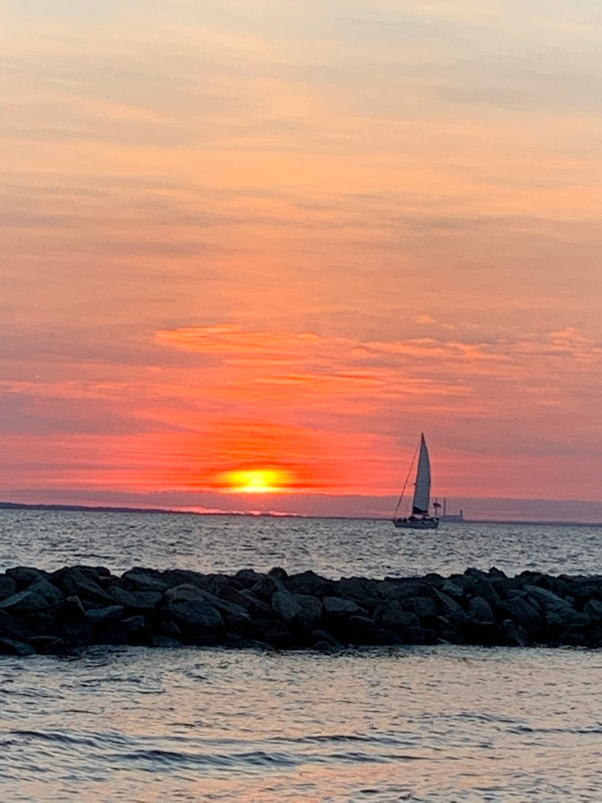 A serene view of a sunset over the water is captured, with vibrant orange and pink hues filling the sky. A sailboat glides across the horizon, silhouetted against the glowing sun, while rocky formations frame the scene in the foreground.