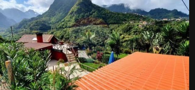 The image presents a panoramic view of lush, green mountains under a partially cloudy sky. A vibrant orange roof is visible in the foreground, with a landscaped garden featuring various plants and trees in the surrounding area. A rustic structure with a terrace can be seen in the mid-ground.