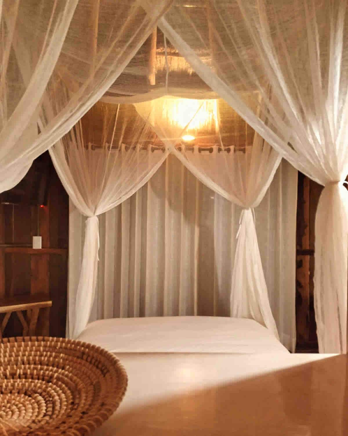 A spacious bed is framed by flowing sheer curtains, creating a serene sleeping area. A warm light fixture illuminates the space, while a woven basket is positioned in the foreground, adding a natural texture to the overall decor.