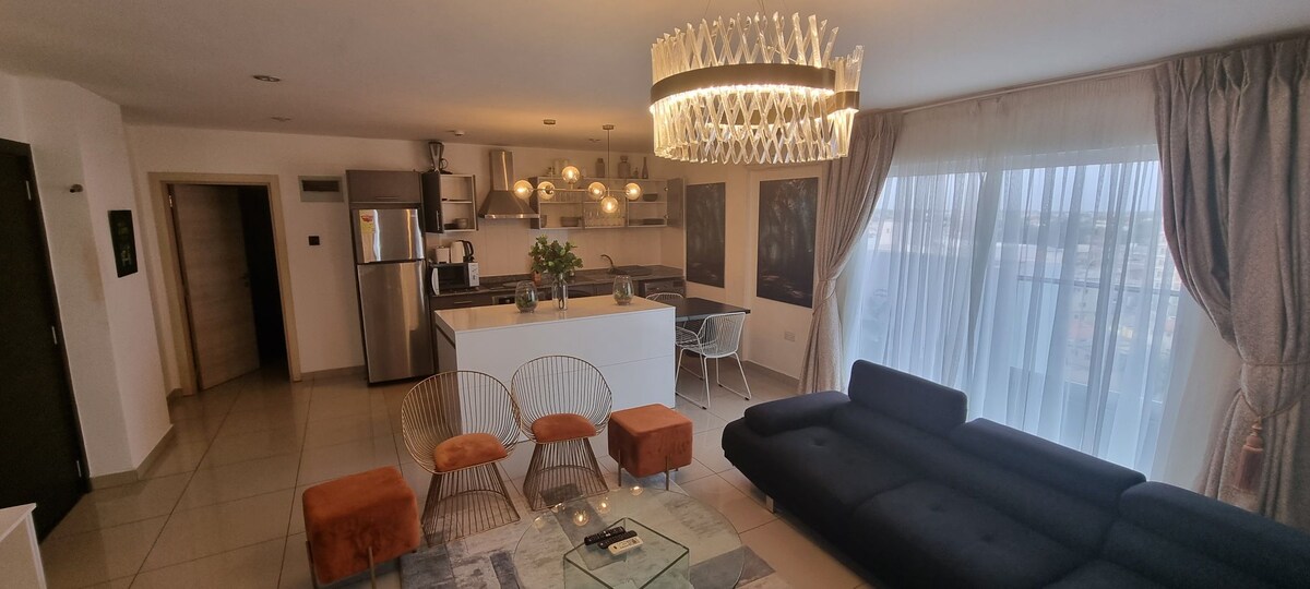 The spacious living area is adorned with modern furnishings, including a sleek dark sofa and three accent chairs. A glass coffee table sits on a textured rug, complemented by a chic chandelier and large windows draped with sheer curtains, allowing natural light to brighten the room.