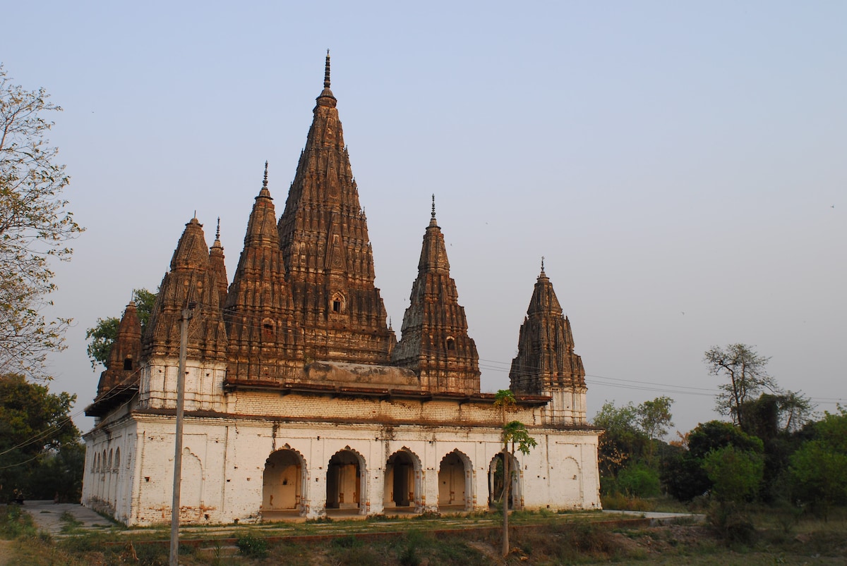 An ancient temple structure showcases distinctive architecture with multiple spires reaching towards the sky. The building features a weathered facade, with decorative elements visible above arched doorways. Surrounding greenery complements the temple, creating a serene atmosphere within the historic landscape.