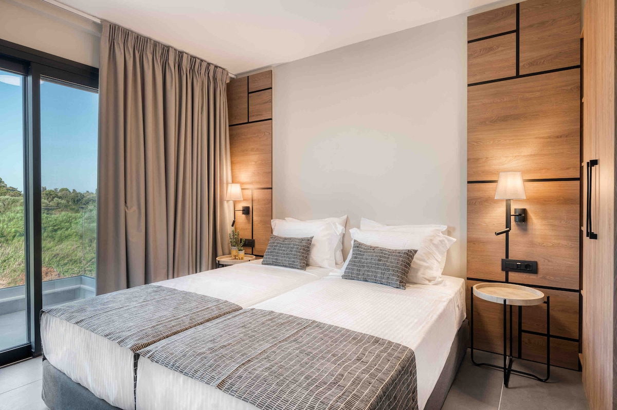 A serene bedroom features two spacious twin beds with light linens, accented by textured gray blankets. Natural light illuminates the room through large glass doors that open to greenery outside, while subtle wood paneling adds a modern touch.