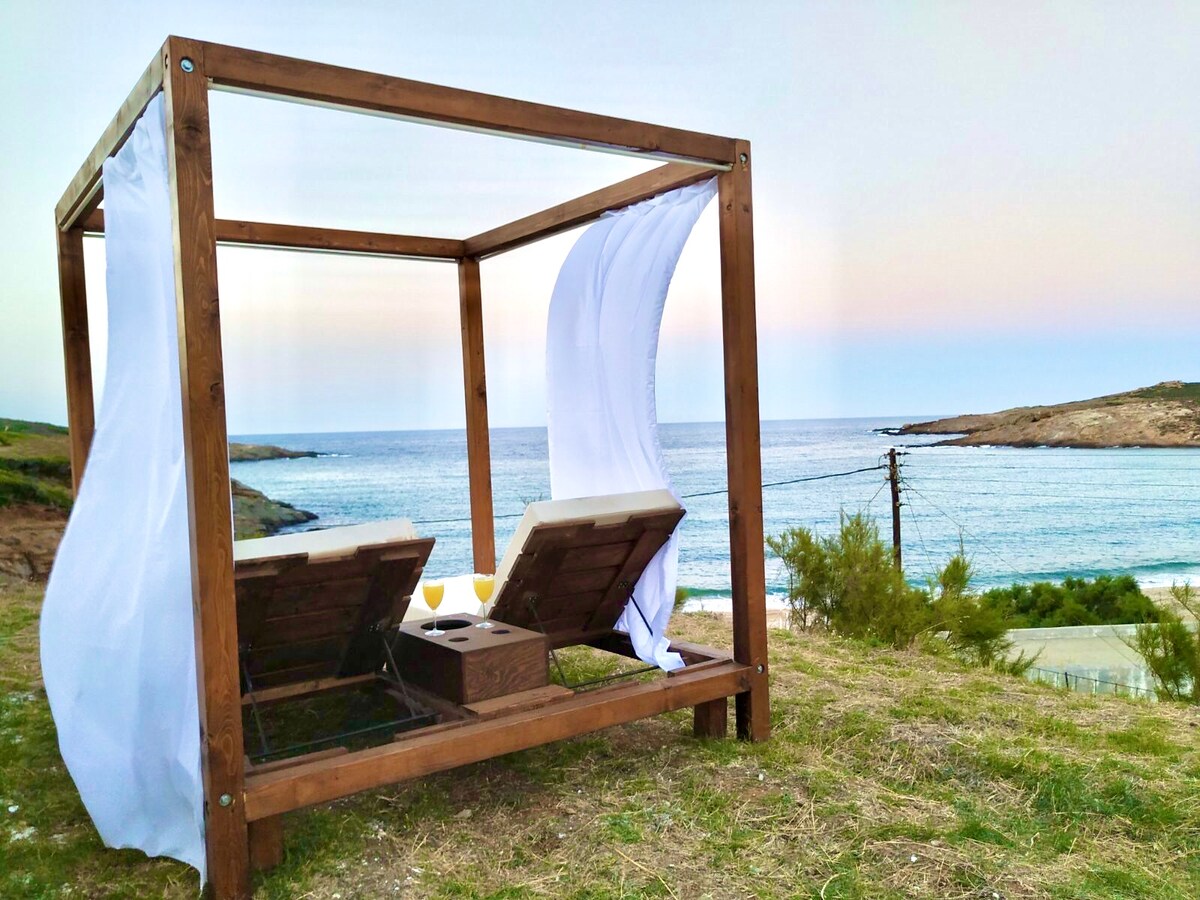 A wooden cabana is situated on a grassy area overlooking the ocean. Two lounge chairs are positioned inside the structure, with soft white drapes gently flowing in the breeze. The scenic view includes calm waves and a soft gradient of colors in the sky.