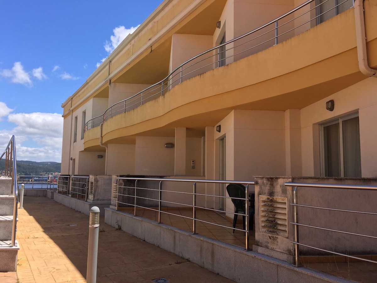 A spacious outdoor terrace is outlined by modern railings, leading to an entrance of a well-designed residential building. Large doors to the interior are visible, with a single chair positioned nearby, offering an inviting spot to enjoy the surroundings. The sky displays scattered clouds.