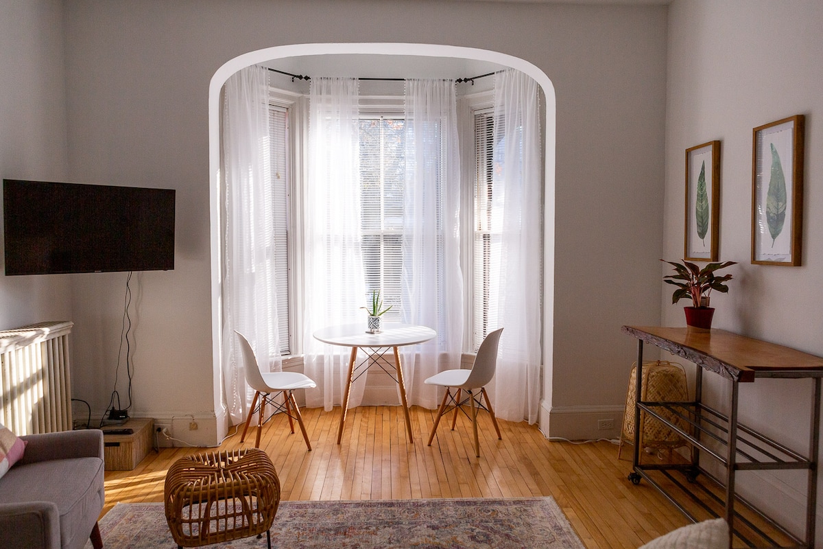 A bright living area features an elegant bay window dressed with sheer curtains, illuminating a small dining table surrounded by two modern chairs. A wall-mounted television, a plant on the table, and framed artwork enhance the space's charm.
