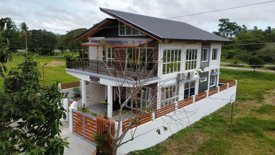 A modern two-story house is showcased, featuring large windows that provide ample natural light. A spacious balcony is visible on the upper level, surrounded by greenery. The property is enclosed with wooden fencing, blending harmoniously with the surrounding landscape.
