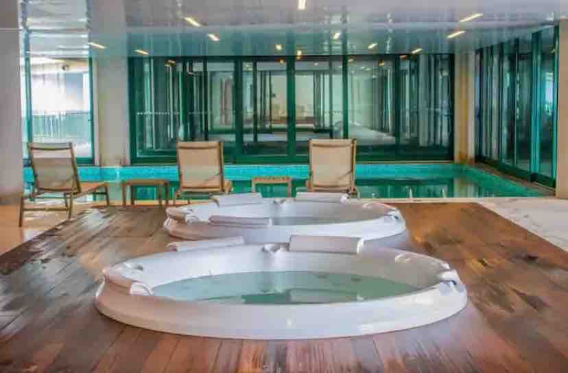 Two jacuzzi tubs are positioned on a wooden platform, surrounded by a spacious indoor area. Behind them, large glass windows provide views of an inviting swimming pool. Several cream-colored lounge chairs are arranged nearby, complementing the serene setting.